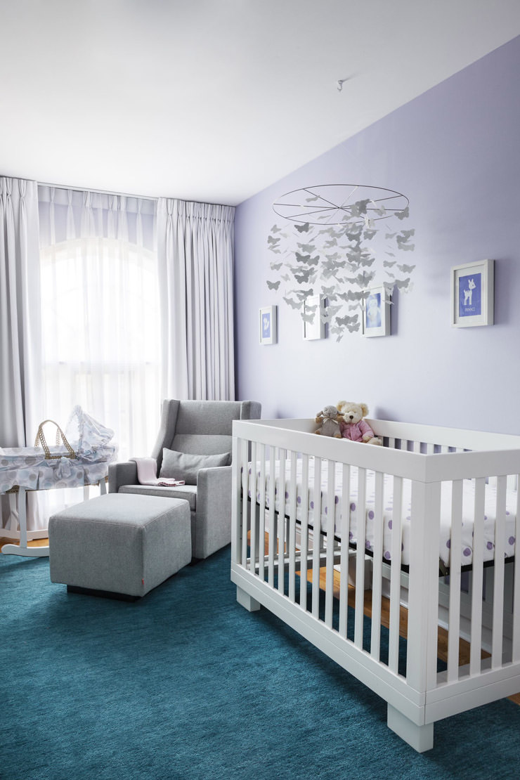 اتاق خواب نوزاد شیک و زیبا با دیوارهای بنفش پاستلی، فرش آبی، مبل شیردهی طوسی و تخت نوزاد سفید که آویز تخت پروانه ای دارد